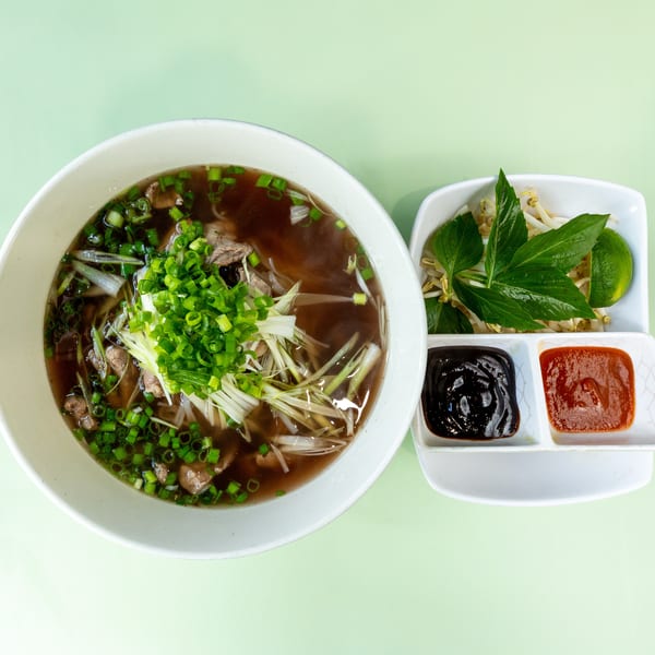A bowl of pho with beef, noodles, and green onions, served with a side of sauces and garnishes.