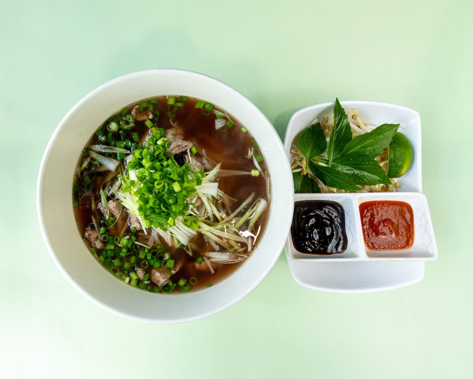 A bowl of pho with beef, noodles, and green onions, served with a side of sauces and garnishes.