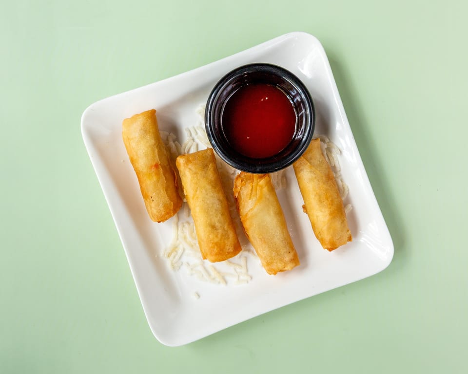 Four golden-brown spring rolls with a side of red dipping sauce on a white rectangular plate.