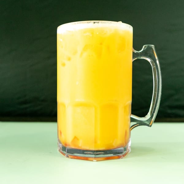 A large clear mug filled with a frothy orange drink, possibly a mango lassi, on a light green surface.