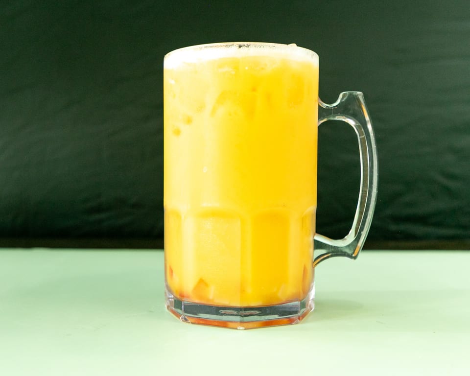 A large clear mug filled with a frothy orange drink, possibly a mango lassi, on a light green surface.