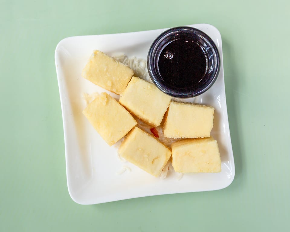 Six rectangular pieces of fried tofu with a small bowl of dark dipping sauce on a white plate.