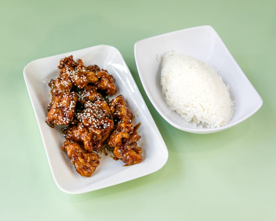 Sweet and sour chicken with sesame seeds and a side of white rice on a light green background.