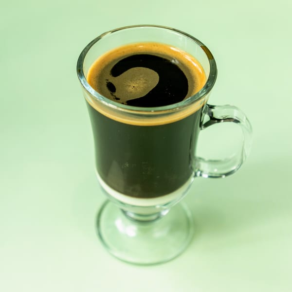 A clear glass mug of dark coffee with a layer of white cream at the bottom.