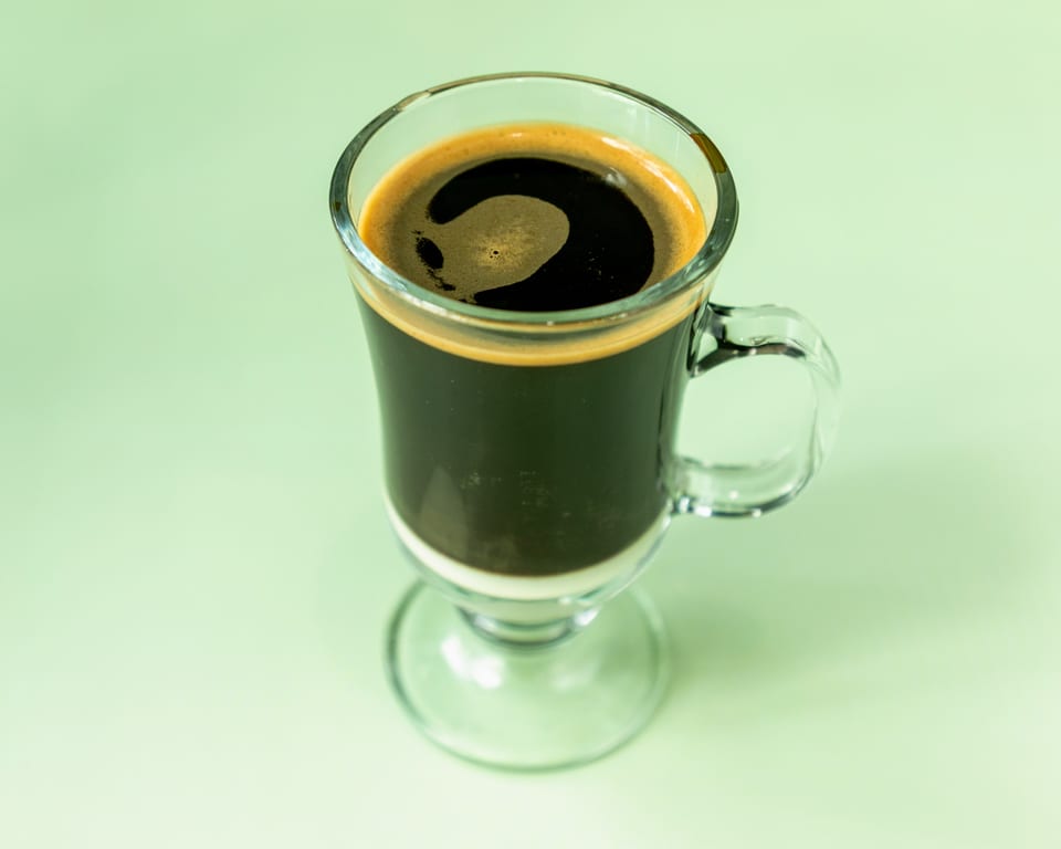 A clear glass mug of dark coffee with a layer of white cream at the bottom.