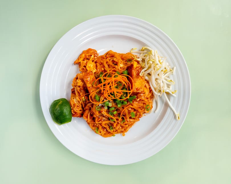 Pad Thai noodles with chicken, bean sprouts, shredded carrots, green onions, and a lime wedge.