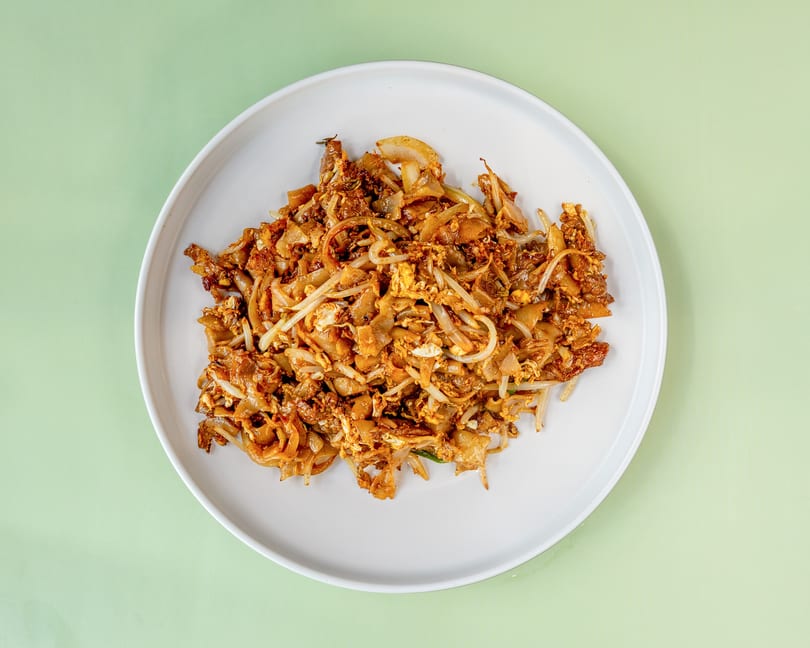 A white plate filled with Char Kway Teow, a stir-fried noodle dish with bean sprouts and egg.