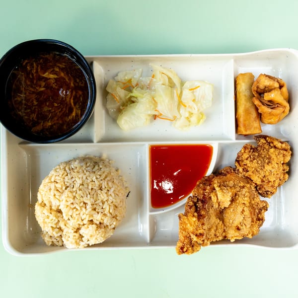 A bento box with fried chicken, rice, soup, pickled cabbage, and spring rolls.