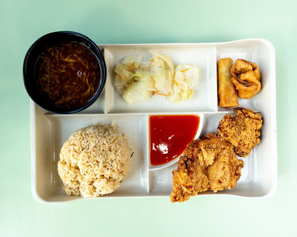 A bento box with fried chicken, rice, soup, pickled cabbage, and spring rolls.