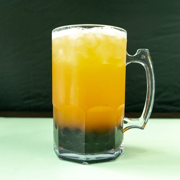 A large clear mug filled with a layered orange and dark brown drink, topped with foam and ice.