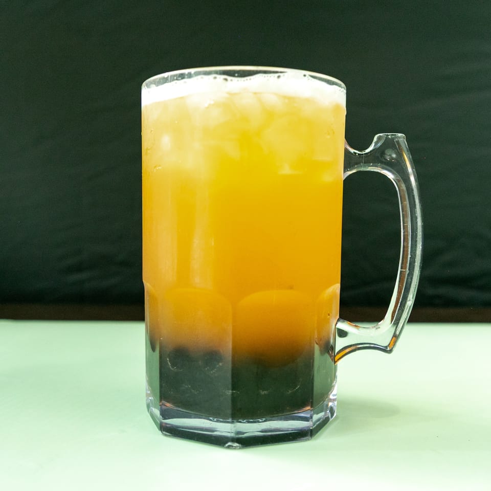 A large clear mug filled with a layered orange and dark brown drink, topped with foam and ice.