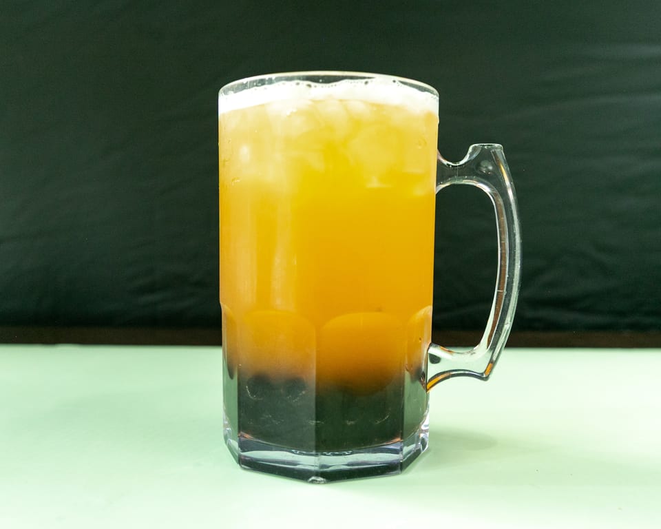 A large clear mug filled with a layered orange and dark brown drink, topped with foam and ice.